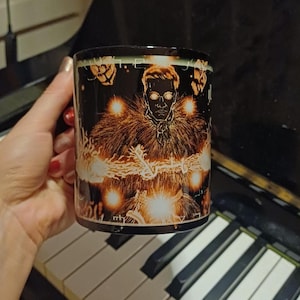 May include: Black coffee mug featuring a detailed illustration of a figure with glowing eyes and a fur-like collar, surrounded by bright, radiating light. The mug has a dark background with additional glowing accents.