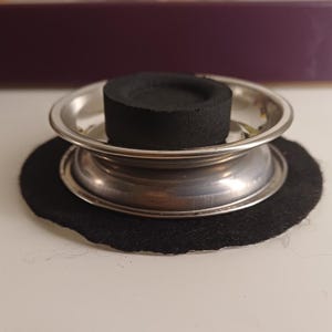 Puede incluir: Un quemador de incienso plateado con un disco de carbón negro, colocado sobre una almohadilla de fieltro negro. El quemador tiene un diseño de dos niveles. El fondo es de color púrpura oscuro.