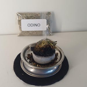 Op de afbeelding: Een aangestoken houtskool wierookschijf met kruiden en rook, in een zilveren metalen wierookhouder op een zwarte onderzetter. Een kleine zak met gedroogde kruiden met het woord "ODINO" gedrukt op een wit etiket staat op de achtergrond.