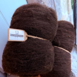 May include: Two bundles of dark brown wool roving with a fuzzy, textured appearance. A small, rectangular tan paper tag is attached to the larger bundle with twine, featuring the printed text The Yorkshire Wool Co. in black ink.