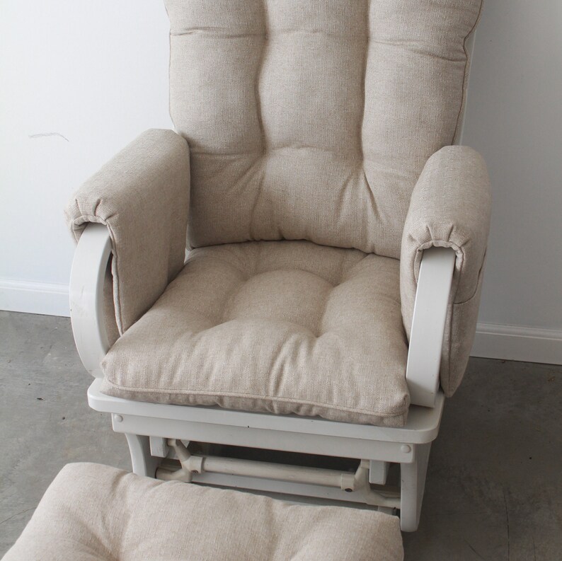 May include: A white rocking chair with a beige fabric cushion and a matching footrest. The chair has a curved back and armrests.