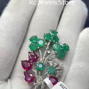 Puede incluir: Broche floral con esmeraldas verdes y rubíes rojos engastados en metal plateado. El broche tiene un diseño detallado con pequeños acentos brillantes. El texto "Rc Jewels Store" es visible en el fondo.