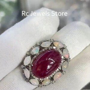 May include: An oval ring with a central, deep red gemstone, encircled by smaller opalescent stones and tiny clear gems. The ring is set in silver-toned metal. The text "Rc Jewels Store" is visible.