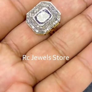May include: A silver and gold ring featuring a rectangular, off-white central stone, encircled by smaller, clear stones. The ring showcases a geometric design and is set on a gold band. The text "Rc Jewels Store" is visible.