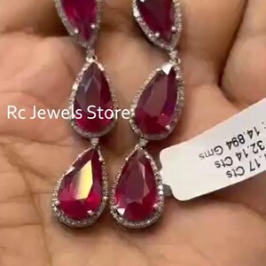 May include: A pair of drop earrings featuring three pear-shaped red gemstones each, surrounded by small white stones. A white tag is attached with the text Rc Jewels Store, 32.14 Cts, and 14.894 Gms. The design is symmetrical and elongated.