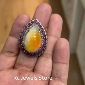 Op de afbeelding: Een druppelvormige ring met een centrale iriserende opaal met gele en oranje flitsen, omringd door een halo van kleine rode edelstenen en een rand van heldere stenen, met de tekst Rc Jewels Store zichtbaar op het onderste gedeelte.