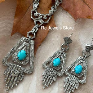 May include: A silver-toned jewelry set featuring a necklace and earrings. The necklace and earrings have a geometric design with a large, oval turquoise stone in the center. The jewelry is encrusted with small, clear stones. The text "Rc Jewels Store" is visible.