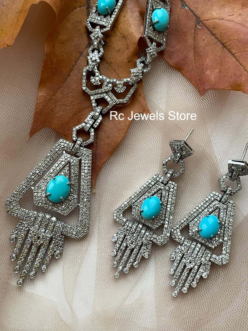 May include: A silver-toned jewelry set featuring a necklace and earrings. The necklace and earrings have a geometric design with a large, oval turquoise stone in the center. The jewelry is encrusted with small, clear stones. The text "Rc Jewels Store" is visible.