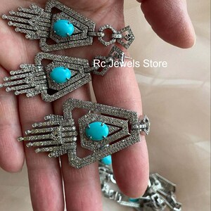 May include: Silver-toned earrings with a geometric design, featuring a central oval turquoise stone. The earrings have a detailed, textured surface and are embellished with small, sparkling accents. The text "Rc Jewels Store" is visible.