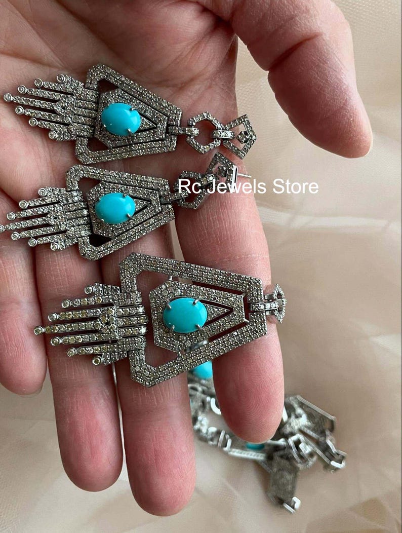 May include: Silver-toned earrings with a geometric design, featuring a central oval turquoise stone. The earrings have a detailed, textured surface and are embellished with small, sparkling accents. The text "Rc Jewels Store" is visible.