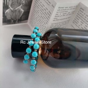 May include: Two turquoise beaded bracelets are displayed on a dark bottle. The bracelets feature round, light blue beads and silver-colored metal. The text "Rc Jewels Store" is visible on the bottle.