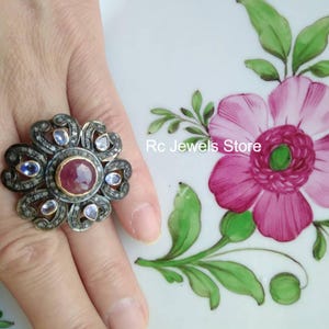 May include: A floral-shaped ring with a large, round, red gemstone at its center. The ring is surrounded by smaller blue and clear gemstones, set in a dark metal. The text "Rc Jewels Store" is visible.