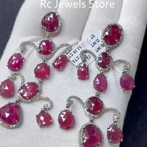 May include: Elegant ruby and diamond earrings. The earrings feature multiple teardrop and oval-shaped rubies, each surrounded by a halo of small diamonds. The design includes curved, diamond-studded accents. The earrings are displayed on a white surface.
