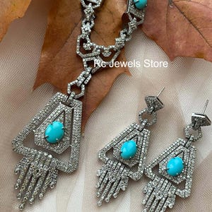 May include: A silver-toned jewelry set featuring a necklace and earrings. The necklace and earrings have a geometric design with a large, oval turquoise stone in the center. The jewelry is encrusted with small, clear stones. The text "Rc Jewels Store" is visible.