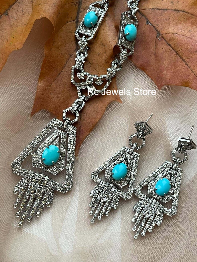 May include: A silver-toned jewelry set featuring a necklace and earrings. The necklace and earrings have a geometric design with a large, oval turquoise stone in the center. The jewelry is encrusted with small, clear stones. The text "Rc Jewels Store" is visible.