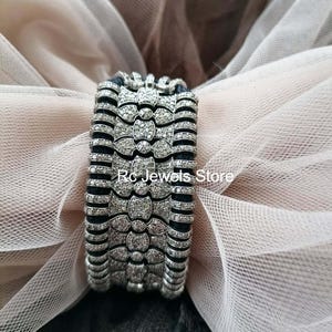 May include: A wide, silver-toned bracelet with a repeating pattern of diamond-like embellishments. The bracelet has a dark, possibly black, backing. The text "Rc Jewels Store" is visible on the image.