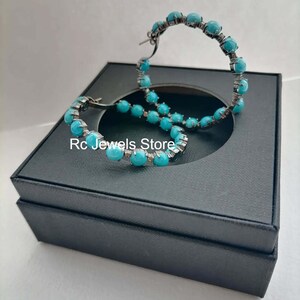 May include: A pair of hoop earrings with turquoise-colored beads. The earrings are set in a dark metal and are displayed on a black jewelry box. The text "Rc Jewels Store" is visible.