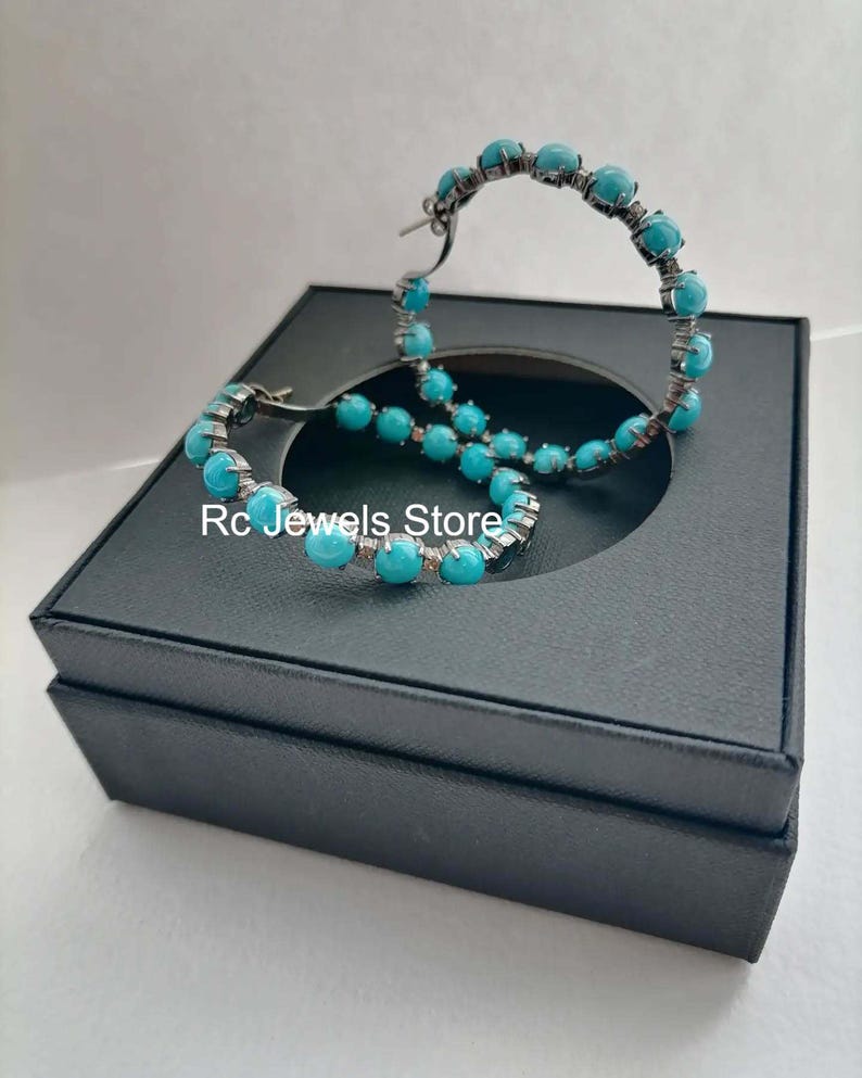 May include: A pair of hoop earrings with turquoise-colored beads. The earrings are set in a dark metal and are displayed on a black jewelry box. The text "Rc Jewels Store" is visible.