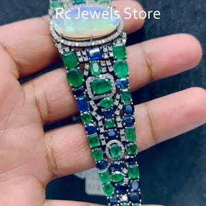 May include: A luxury bracelet with an oval opal at the top, encircled by diamonds. The bracelet is decorated with emeralds, sapphires, and diamonds, creating a vibrant colour contrast. The text "RC Jewels Store" is visible.