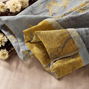 May include: Close-up of folded fabric with a gray and gold floral pattern. The fabric has a textured appearance and is folded to reveal the contrasting colors. The edges are trimmed with a darker gray border. Dried flowers are in the background.