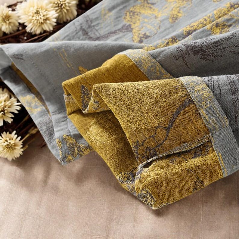 May include: Close-up of folded fabric with a gray and gold floral pattern. The fabric has a textured appearance and is folded to reveal the contrasting colors. The edges are trimmed with a darker gray border. Dried flowers are in the background.