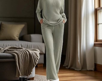 May include: A sage green ribbed knit pajama set. The set includes a long-sleeved V-neck top and matching wide-leg pants. The person is wearing white fuzzy slippers. The outfit is made of a soft, comfortable material.