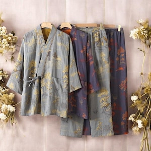 May include: Three-piece pajama set in a soft, muted color palette. The set includes a kimono-style top with a tie closure, and two pairs of pants. The fabric features a repeating pattern of trees and buildings in shades of gold, red, and blue.