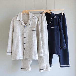May include: Three sets of pajamas hanging on a wooden rod. One set is light gray with black trim, another is light gray, and the third is navy blue with white trim. Each set includes a button-down shirt and pants.