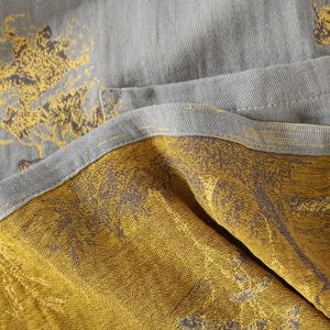 May include: Close-up of a folded textile with a tree pattern. The fabric is a muted gray on one side and a mustard yellow on the other, with the tree design in a darker shade. The edges are neatly sewn.