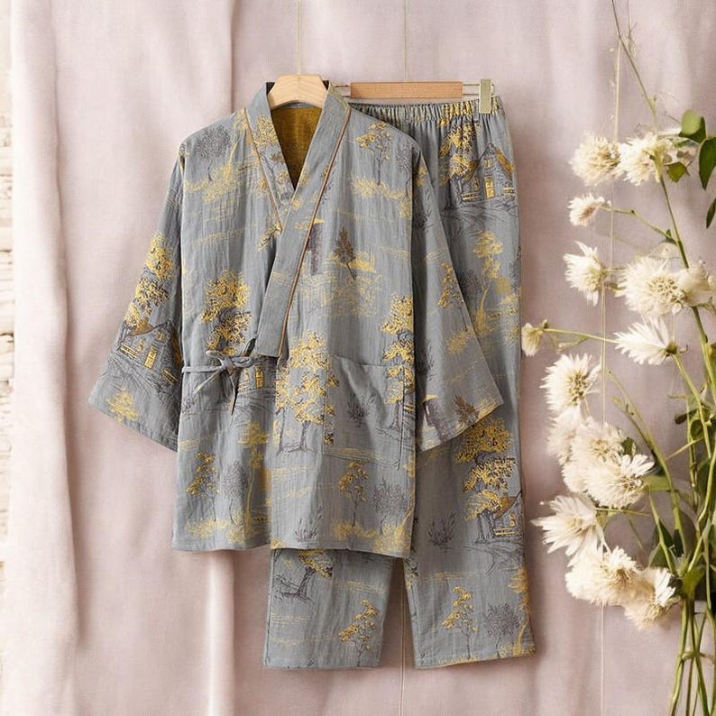 May include: A gray pajama set with a matching top and pants. The set features a landscape design in gold and gray tones. The top has a wrap-style closure with a tie, and the pants have an elastic waistband. The set is hanging on a wooden hanger.