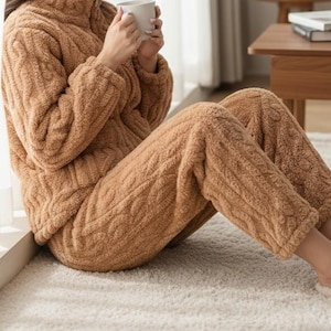 May include: A cozy, brown, cable-knit pajama set. The set includes a zip-up jacket with a high collar and matching pants. The person is holding a white mug. The pajamas appear soft and comfortable, perfect for lounging.