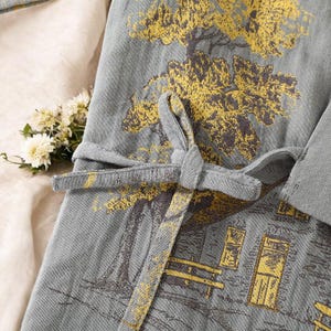 May include: Close-up of a gray robe with a yellow and brown tree and building print. The robe has a tie closure and a soft, textured appearance. A small bouquet of white flowers rests on the fabric.