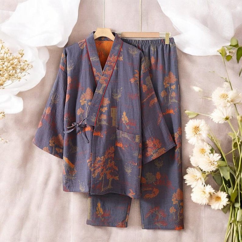 May include: A two-piece pajama set in a muted blue hue, adorned with a repeating pattern of orange and brown trees and foliage. The set includes a kimono-style top with a tie closure and matching pants. The top has a red trim and a pocket.