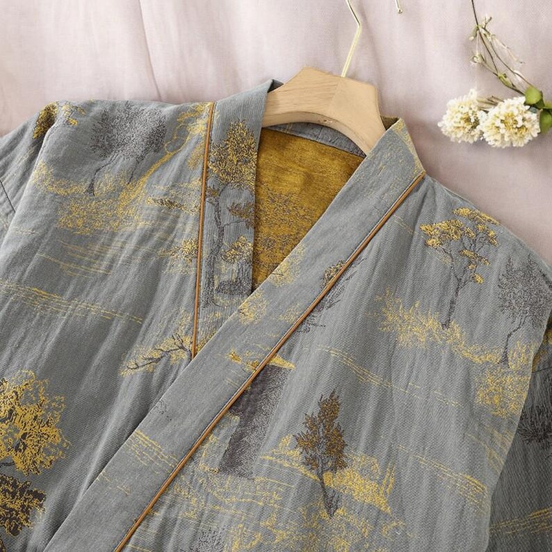 May include: A gray kimono-style robe with a landscape design featuring gold-colored trees and accents. The robe has a contrasting gold interior and brown trim along the edges. It is hanging on a wooden hanger.