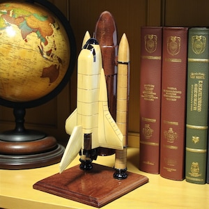 May include: A wooden model of a space shuttle, painted in shades of tan, brown, and black, is displayed on a wooden base. The model is positioned in front of a globe and several books with maroon and green covers.