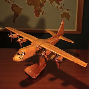 May include: A detailed wooden model of a four-engine aircraft, with a light brown finish. The model has four propellers and a detailed cockpit. It is displayed on a wooden stand. A framed world map is visible in the background.
