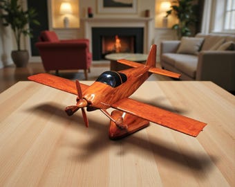 Könnte beinhalten: Ein Holzmodellflugzeug mit einem dunkelbraunen Propeller, Flügeln und Rumpf. Das Modell hat ein schwarzes Cockpit und ist auf einem Holzständer montiert. Das Flugzeug steht auf einem hellbraunen Holztisch.