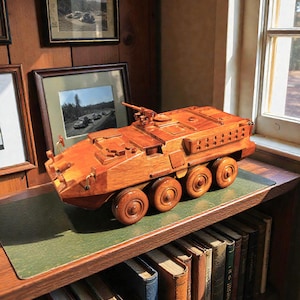 Puede incluir: Maqueta de madera detallada de un vehículo blindado, con ocho ruedas y una torreta. El modelo está hecho de madera marrón rojiza y se encuentra en una estantería con una superficie verde, rodeado de libros y fotos enmarcadas.
