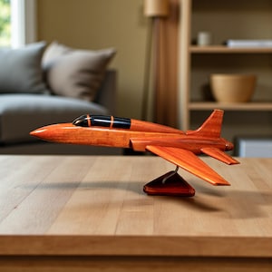 May include: A wooden model airplane with a sleek, orange finish and black cockpit. The model is displayed on a wooden table, showcasing its detailed design and craftsmanship. The wings and tail are visible, highlighting the aerodynamic shape.