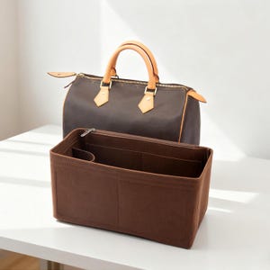 May include: A brown handbag with tan leather handles and trim, and a matching brown felt organizer. The handbag has a zipper closure and gold-toned hardware. The organizer has multiple compartments.