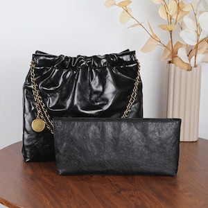 May include: Black quilted leather handbag with a gold chain strap and a matching black clutch. The handbag has a drawstring closure and a gold medallion charm. The clutch is rectangular and has a textured surface. A vase with decorative branches is in the background.