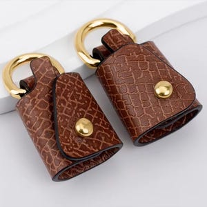 May include: Two brown leather keychains with a textured pattern and gold-colored metal accents. Each keychain has a gold ring and a snap closure. The keychains are displayed against a white background.