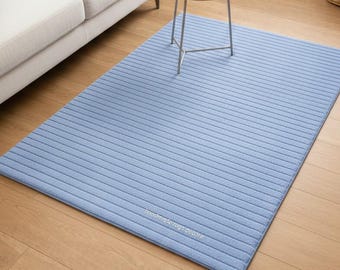 May include: A rectangular light blue area rug featuring a pattern of thin, horizontal, darker blue stripes. The text "handmade rugs decore" is printed in small white letters near the bottom edge of the rug, which lies flat on a wooden floor.