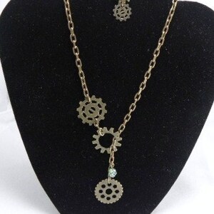 May include: A gold necklace with three gear charms and a small green crystal charm. The necklace is on a black display stand. The necklace also includes a pair of matching earrings with gear charms.