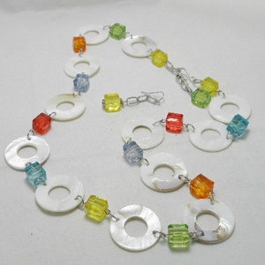 May include: A white shell necklace with colorful beads. The necklace is made of white shell circles connected by silver chain. The beads are in various colors, including blue, green, yellow, orange, and red.