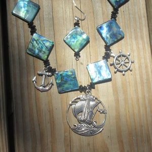 May include: A silver necklace with a ship charm, an anchor charm, and a ship's wheel charm. The necklace is made with black beads and iridescent green and blue abalone shell squares.