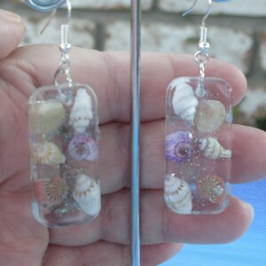 May include: A pair of silver earrings with clear rectangular resin charms. Each charm contains a variety of small seashells and iridescent glitter.
