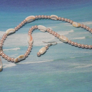 May include: A necklace and earring set featuring a seashell pendant and shell accents. The necklace is strung with small, pink beads and includes a silver-toned clasp. The earrings have a matching design. The background is a blue and white gradient.