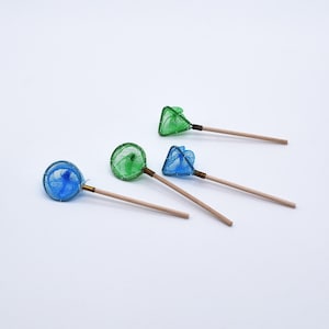May include: Four miniature fishing nets in blue and green with wooden handles. The nets have different shapes, including round and triangular, and are arranged on a white surface. The nets are likely for a dollhouse or other miniature project.