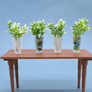 May include: Four miniature decorative plants in clear glass vases filled with white pebbles and colorful beads, arranged on a small wooden table. The plants have green leaves and white flowers, set against a light blue background.